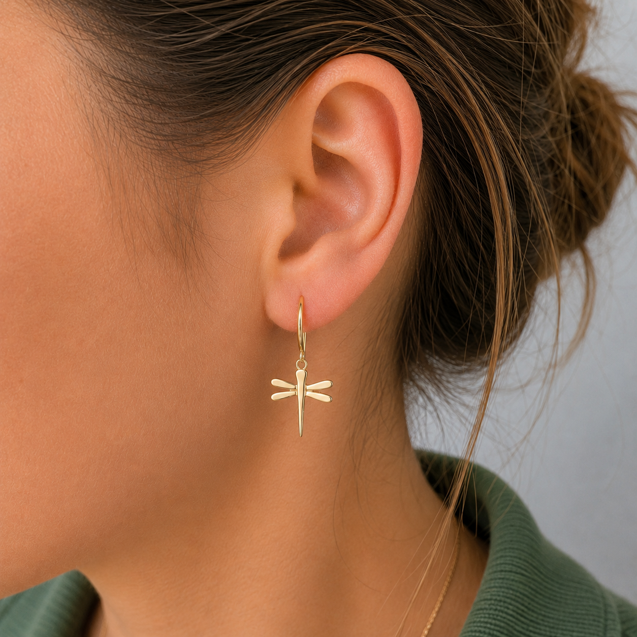 A close-up of a woman's ear wearing a delicate gold dragonfly drop earring, with her hair tied back and a green top visible.